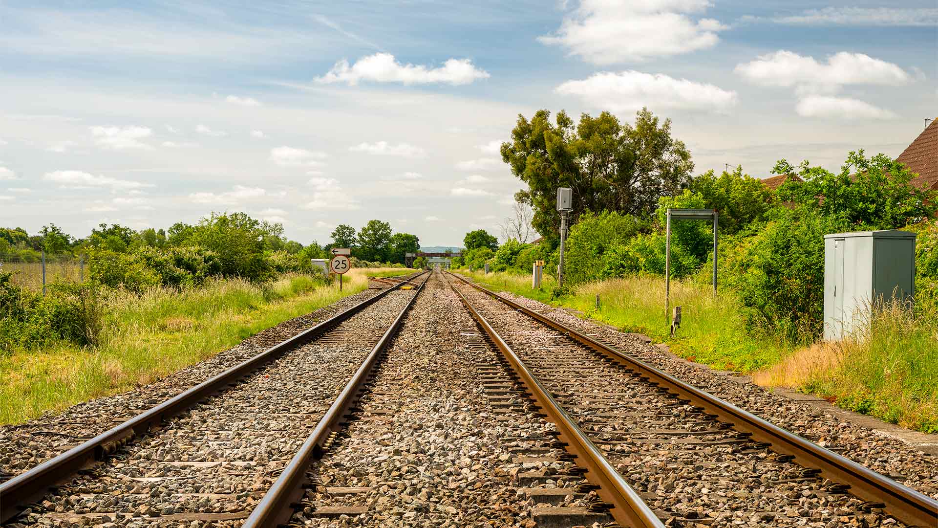 Railway tracks extend through countryside setting with green vegetation, trackside equipment and 25 speed limit sign, under a partly cloudy sky.